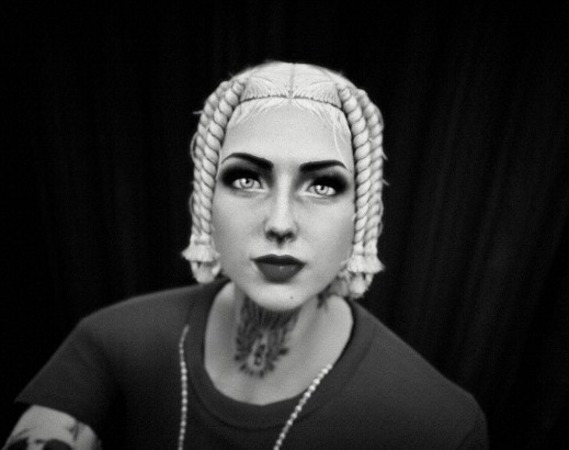 It is a monochromatic (black and white) close-up, photorealistic digital illustration, serving as the cover art for the "AureliaWRLD (Anita Lacey)" Transport Fever mod creator profile on Mod.io. The image is a moody portrait of AureliaWRLD, viewed from a low angle, looking slightly up and directly into the camera with an intense, steady gaze. Her face is centered, and her expression is serious and direct. Her platinum-blonde, braided hair is intricately styled into multiple small cornrows that start at her hairline and curve backward, creating a structured framing effect around her head. Her eyes are dark, intense, and heavily lined, with a prominent white-dot highlight in each, giving her eyes a striking quality. Her lips are full and dark-colored. Extensive, complex black-ink tattoos completely cover her neck and throat, with intricate, symmetrical patterns that wrap around her. Another portion of a tattoo is visible on her left arm in the lower foreground, though partially obscured. She is wearing a simple, dark-colored t-shirt. A textured, multi-link chain necklace hangs around her neck, with some elements visible below her throat. The background is a deeply textured, pitch-black fabric, providing a clean, dramatic negative space to isolate the subject. The lighting is focused and dramatic, highlighting the contours of her face, the details of her hair and tattoos, and the texture of her clothing. A small, blue square Mod.io logo is visible as an overlay in the upper central portion of her forehead, as a unique branding element for the cover art. The overall aesthetic is similar to a high-end, contemporary artist or musician's album cover, combining classical portraiture with modern elements.